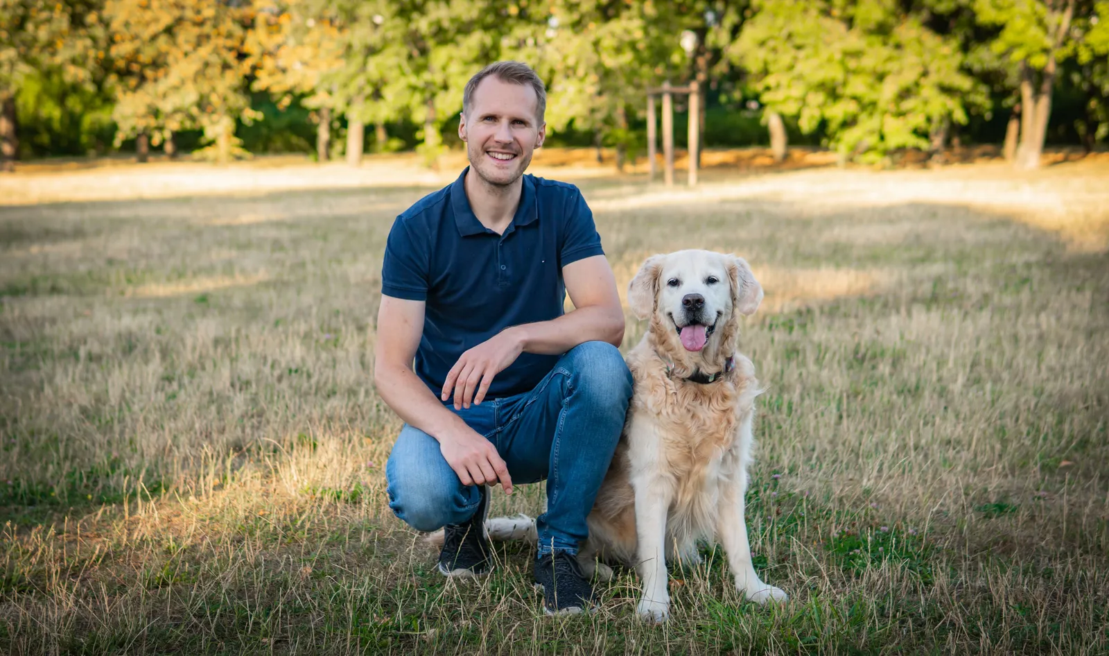 Michał z Petsy kuca na trawie w parku obok złotego retrievera
